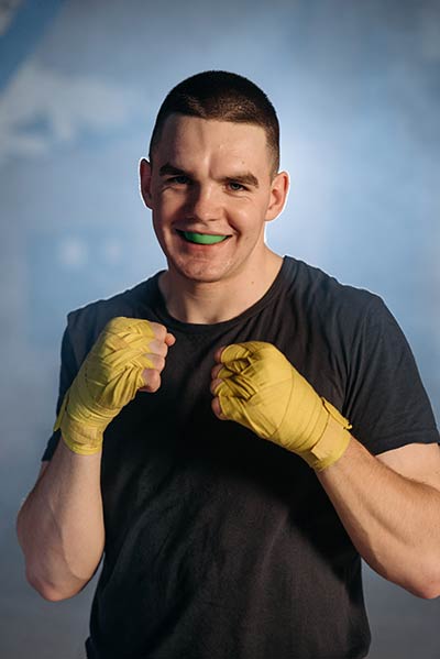A boxer wearing some yellow hand wraps and a green mouthguard poses for the camera.